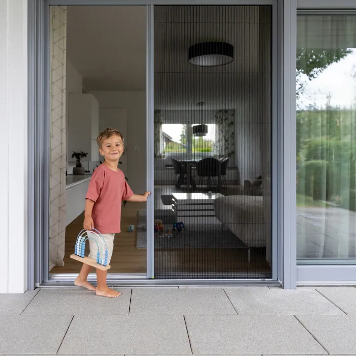 Kind steht vor einer geöffneten Terrassenschiebetür mit Fliegengitter in einem modernen Wohnraum mit Blick nach draußen